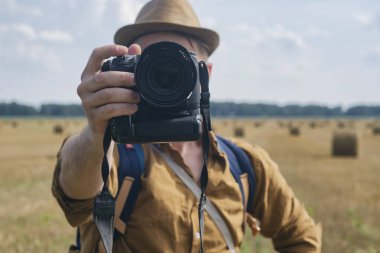 Gezgin fotoğrafçı elinde kamerayla bir tarlanın arka planına ve samanlıklara bakıyor.