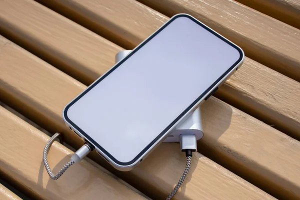 公園内の木製のベンチに白い画面でモックアップスマートフォンを充電パワーバンク — ストック写真