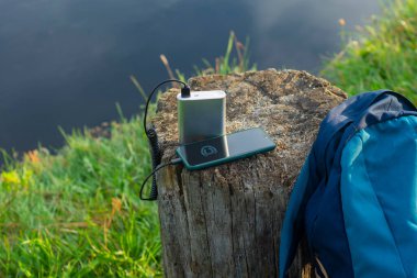Akıllı telefon taşınabilir şarj aleti ile şarj edilir. Elektrik Bankası açık havada telefon faturasını doğa ve nehir arka planında turizm için bir sırt çantasıyla ödüyor.