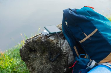 Akıllı telefon taşınabilir şarj aleti ile şarj edilir. Elektrik Bankası açık havada telefon faturasını doğa ve nehir arka planında turizm için bir sırt çantasıyla ödüyor.