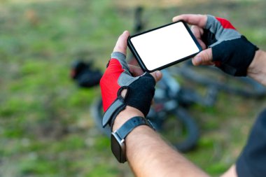 Bisikletçi, doğadaki bir bisikletin arka planında beyaz bir ekranla akıllı telefonu yakın plan çeker.