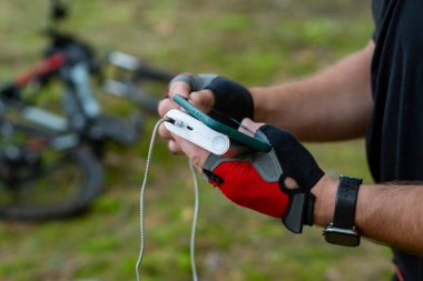 Ormandaki bir bisikletin arka planında elektrik bankasıyla akıllı telefonu şarj eden bir adam.