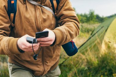 Adam elinde bir akıllı telefon tutuyor ve onu bir enerji bankasıyla turist çadırının arka planına karşı dolduruyor. Taşınabilir seyahat şarj cihazı