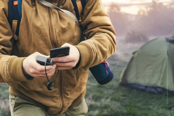 인간은 스마트폰을 자연계에 텐트의 배경에 대항해 은행으로 휴대용 여행용 충전기 — 스톡 사진
