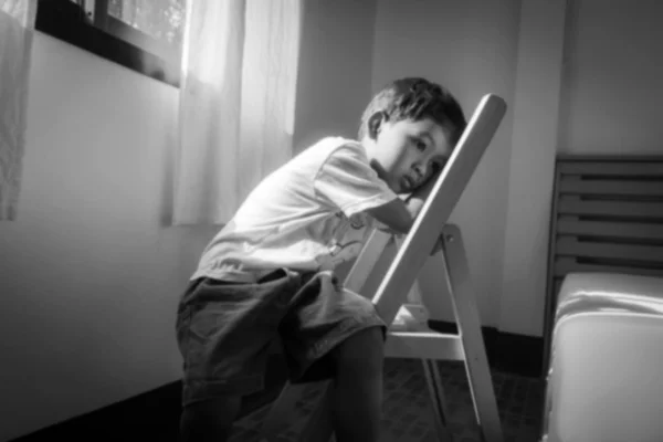 เบลอของเด็กน้อยนั่งคนเดียวบนเก้าอี้ที่หน้าต่างในห้องเบลอ — ภาพถ่ายสต็อก