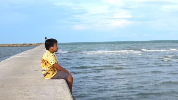 Netter kleiner Junge sitzt allein auf dem Gehweg am Meer — Stockvideo