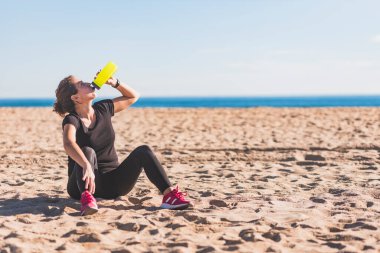 Orta yaşlı bir kadın sahilde egzersiz yaptıktan sonra su içiyor.