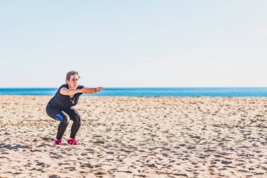 Orta yaşlı bir kadın sahilde egzersiz yapıyor.