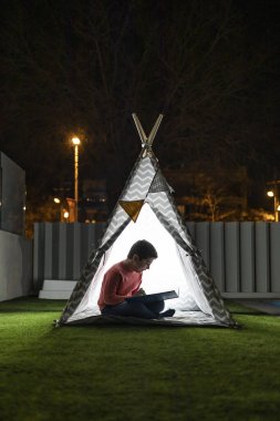 Küçük çocuk geceleri Kızılderili çadırında kitap okuyor.