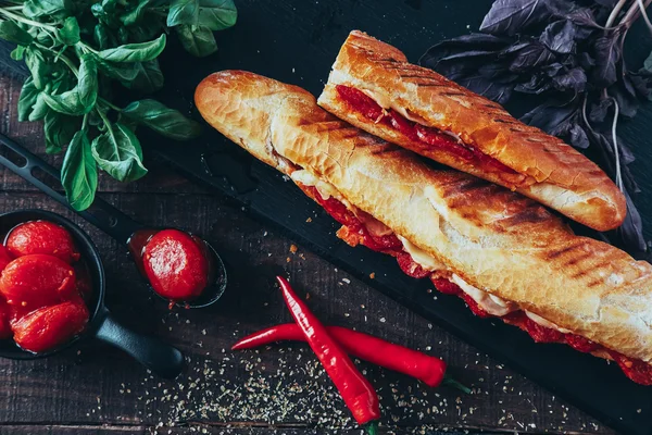Long baguette sandwich with lettuce, vegetables, salami, chili and cheese on black background — Stock Photo, Image