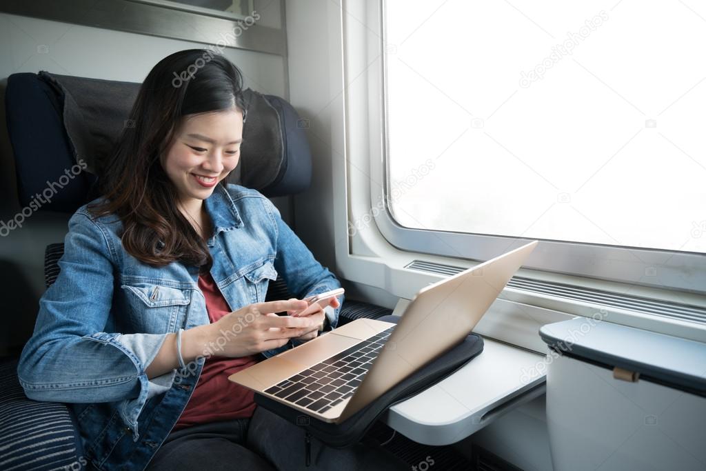 Asian woman smiling at smartphone with laptop on train, copy space on window