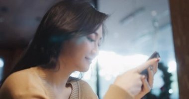 Genç ve güzel Asyalı bir kadın kafeteryada akıllı telefon kullanıyor, mutlu bir gülümseme. İnsanlar modern yaşam tarzı, internet iletişim teknolojisi veya online alışveriş konsepti. El bilgisayarı ağır çekim