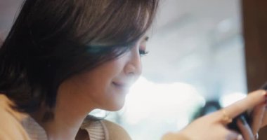 Kafede akıllı telefon kullanan güzel bir Asyalı kadın, mutlu bir gülümseme. İnsan yaşam tarzı, internet iletişim teknolojisi veya online alışveriş konsepti. El bilgisayarı ağır çekim