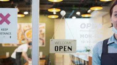 Asyalı kadın restoran sahibinin portresi maske takıyor, kollarını kavuşturuyor ve restoran girişinde ağır çekimde hoş geldin tabelasıyla gülümsüyor. İş, covid-19 koronavirüs salgınından sonra yeniden açıldı