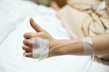 Patient hand thumbs up while given saline IV drip injection on hospital bed clipart