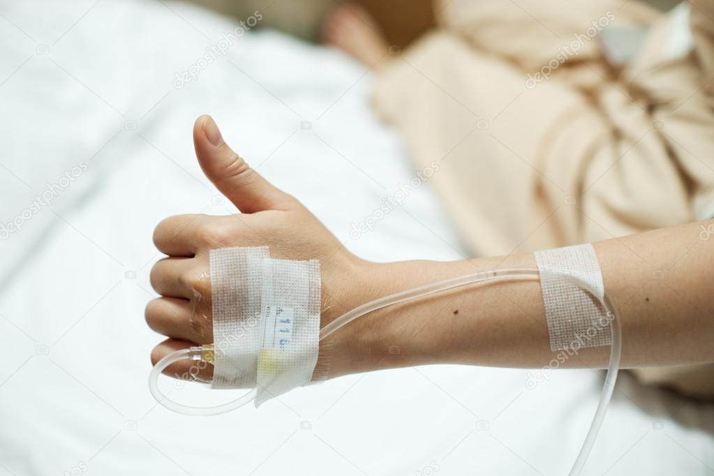 Patient hand thumbs up while given saline IV drip injection on hospital bed