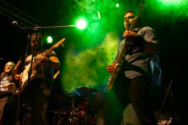 Pair of guitarists playing electric guitars accompanying the drummer and vocalist while the artificial mist is illuminated in green, enveloping the atmosphere in nightlife in Mexico City clipart