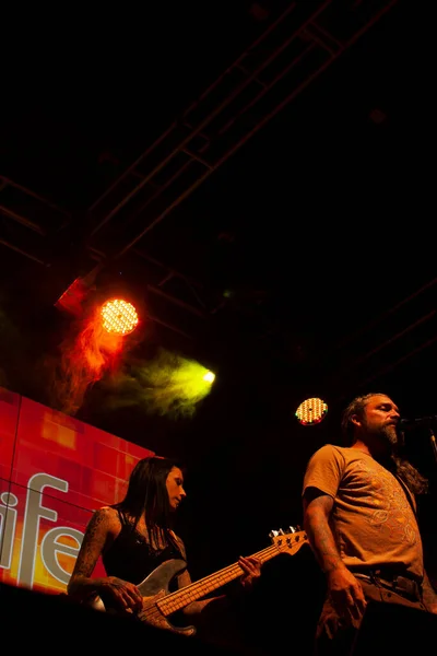 Tattooed and made-up dark woman with smokey eyes playing the bass next to the singer of the band singing illuminated with red and green light surrounded by artificial mist