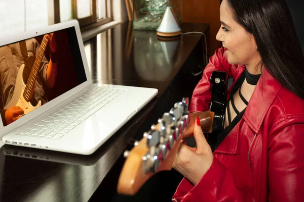 Smiling woman wearing red leather jacket in front of computer attending virtual guitar class in the living room of her home in quarantine