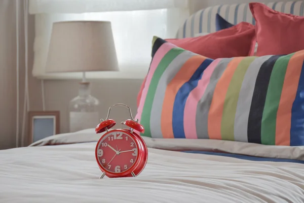 Reloj rojo sobre manta blanca y almohadas a rayas de colores — Foto de Stock