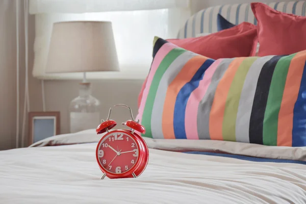 Reloj rojo sobre manta blanca y almohadas a rayas de colores — Foto de Stock