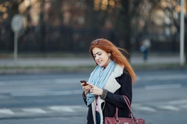 Red-haired girl walking on the street and listen music on her phone