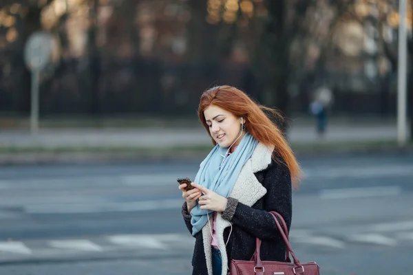 Рыжая девушка ходит по улице и слушает музыку на телефоне — стоковое фото