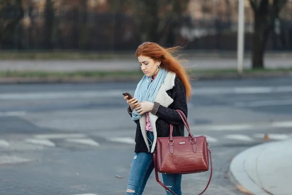 Рыжая девушка ходит по улице и слушает музыку на телефоне — стоковое фото