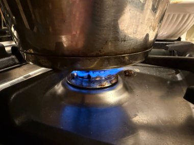 Altında canlı mavi bir alev bulunan bir tencere, gaz sobasının üzerinde oturur. Mutfak ortamında etkin yemek pişirildiğini gösterir..