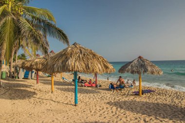Trinidad, Küba - 17 Temmuz 2018: Ziyaretçiler Trinidad Küba 'da kumlu bir sahilde palmiye ağaçlı şemsiyeler altında sıcak Karayip güneşi ve denizin tadını çıkarırken dinleniyorlar.