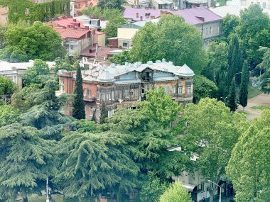 Tiflis, Gürcistan - 27 Nisan 2025: Karmaşık mimarisi olan eski bir bina, Tiflis Gürcistan 'da canlı yeşil yapraklarla çevrili yüksek ağaçların arasına kurulmuştur..
