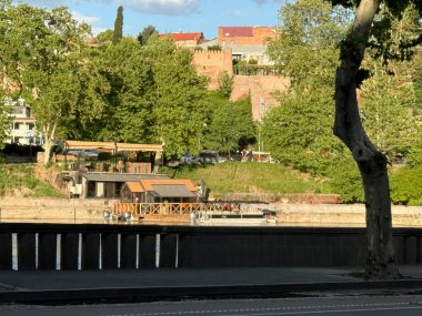 Tiflis, Gürcistan - 27 Nisan 2025: Ziyaretçiler, arka planda yemyeşil ve tarihi binalar bulunan Tiflis 'in nehir kenarında güneşli bir öğleden sonranın keyfini çıkarıyorlar.