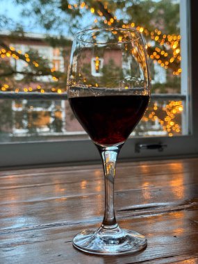 Bir bardak kırmızı şarap, arka planda yanıp sönen ışıklarla dolu ahşap bir masanın üzerinde duruyor ve sıcak bir akşam atmosferi yaratıyor..