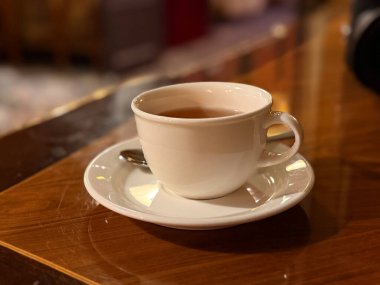 Den Haag, Hollanda - 10 Ekim 2025: İçi çay dolu basit bir beyaz fincan, rahat bir kafe ortamındaki parlak ahşap bir masanın üzerinde duruyor..