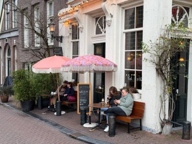 Amsterdam, Hollanda - 15 Ocak 2025: Bir grup arkadaş güneşli bir bahar günü güneşli bir günde içki içerken pembe şemsiyeli rahat bir kafe önünde dinleniyor.