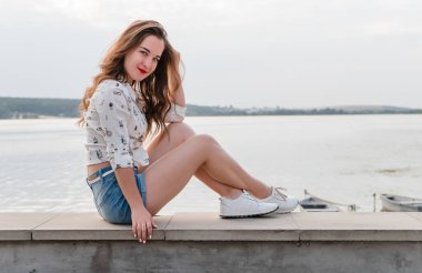 Şortlu ve spor ayakkabılı esmer bir kız büyük bir gölün yanında yürüyor. Şehir gölünün kıyısında kısa kot şortlu bir kız..