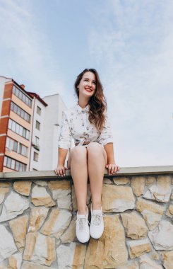 Güzel, gülümseyen bir kız, arka planda taş bir duvarda oturuyor. Uzun saçlı, şortlu, şehirde gezen mutlu kız..