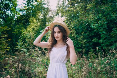 Yeşil yaz ormanlarının arka planında hasır şapkalı genç bir kadın. Tarlada bir buket kurumuş çiçekli elbise giymiş güzel bir kız. Yazlık elbiseli kız..