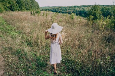 Büyük beyaz şapkalı, buketli bir kadın otların arasında yürüyor. Beyaz elbiseli uzun saçlı kız tarlada koşuyor. Bir yaz akşamı.