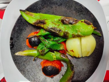 Kömürleşmiş yeşil biberler, kavrulmuş domatesler ve kavrulmuş bir tabakta duran yarım bir soğan, ateşin öptüğü tatların ve mutfak dayanıklılığının ham özünü sembolize ediyor..