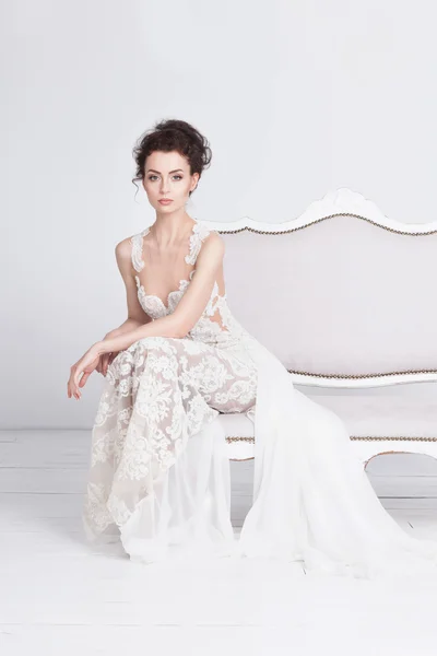 Bella giovane sposa in un lussuoso abito da sposa in pizzo. Si siede su un divano vintage bianco . — Foto Stock