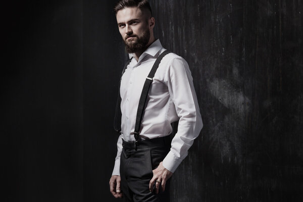Привлекательный бородатый мужчина в белой рубашке и подтяжках, стоящих у темной стены
.