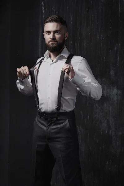 Attractive bearded man in a white shirt and suspenders standing near dark wall.
