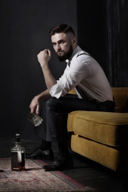 A handsome brutal bearded man sitting on an orange couch. He is holding an empty glass. Whiskey bottle standing on the floor clipart