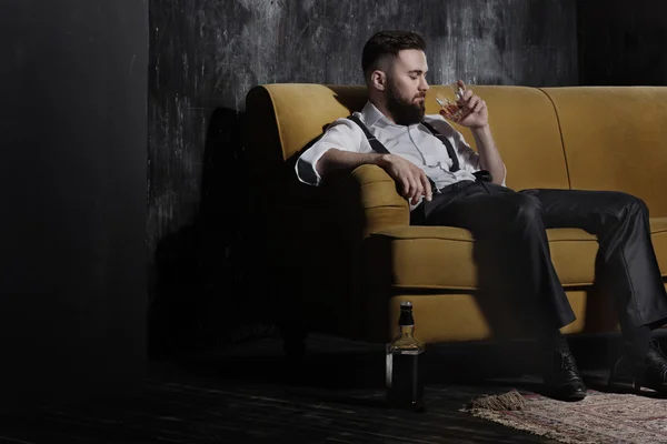 Un guapo barbudo brutal sentado en un sofá naranja. Él está sosteniendo un vaso con un poco de bebida — Foto de Stock