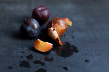 Bir Erik dilim, bir soyulmuş erik ve siyah bir masanın üzerinde yatan bir kaç Bütün Konfit Erik. 