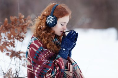 Young red-haired woman drinking a hot drink from a mug in the winter park.
