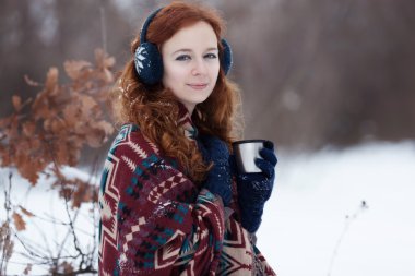 Attractive young red-haired woman drinking a hot drink from a mug in the winter park.