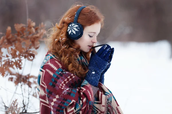 Joven pelirroja bebiendo una bebida caliente de una taza en el parque de invierno . — Foto de Stock