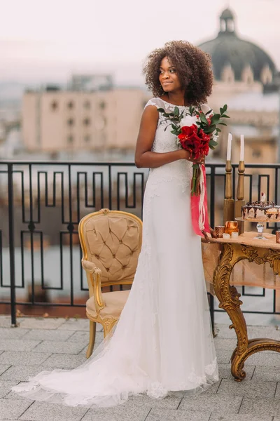 Charming black bride with wedding bouquet in hands standing on the terrace